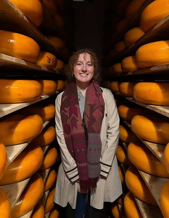 A Converse student stands smiling between massive wheels of cheese at a cheese museum in Amsterdam.